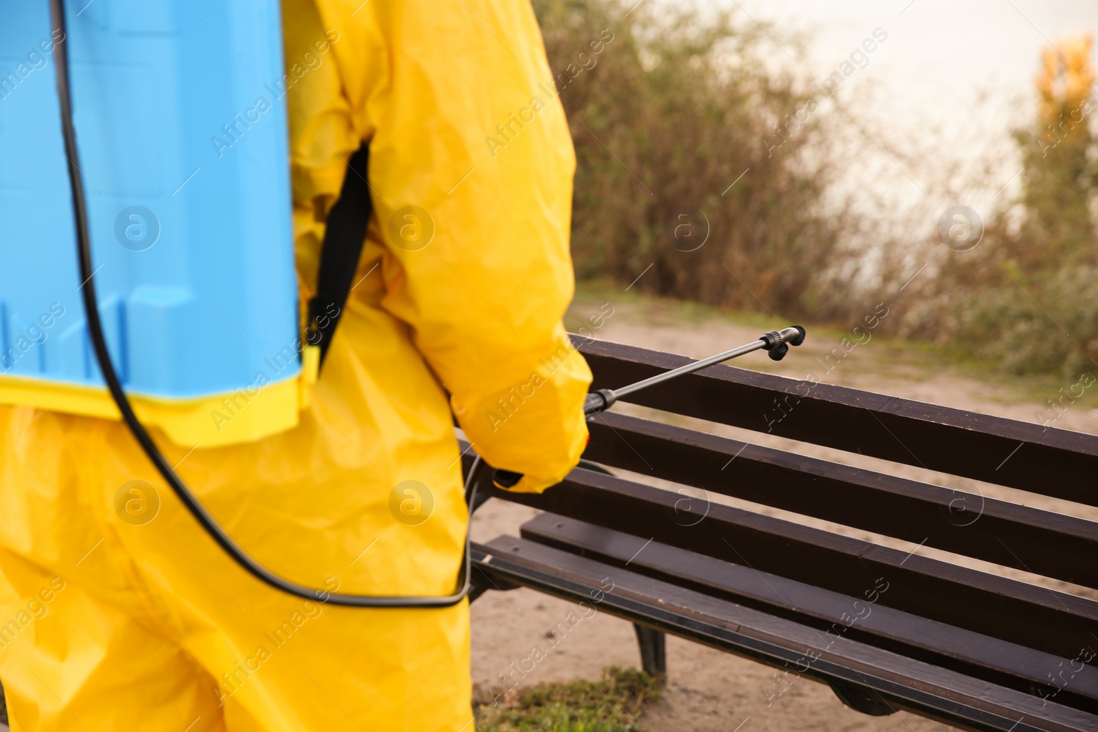 Photo of Person in hazmat suit disinfecting bench in park with sprayer, closeup. Surface treatment during coronavirus pandemic