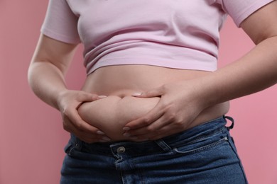 Woman touching belly fat on pink background, closeup. Overweight problem