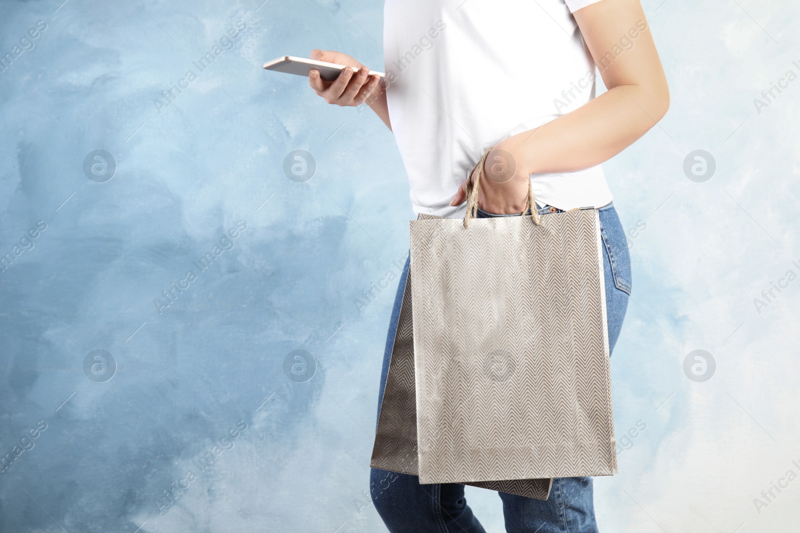 Photo of Woman with silver shopping paper bags and smartphone on light blue background, closeup. Space for text