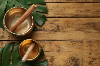 Golden singing bowls, mallets and monstera leaves on wooden table, flat lay. Space for text