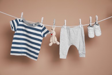 Cute small baby shoes, clothes and toy hanging on washing line against brown background