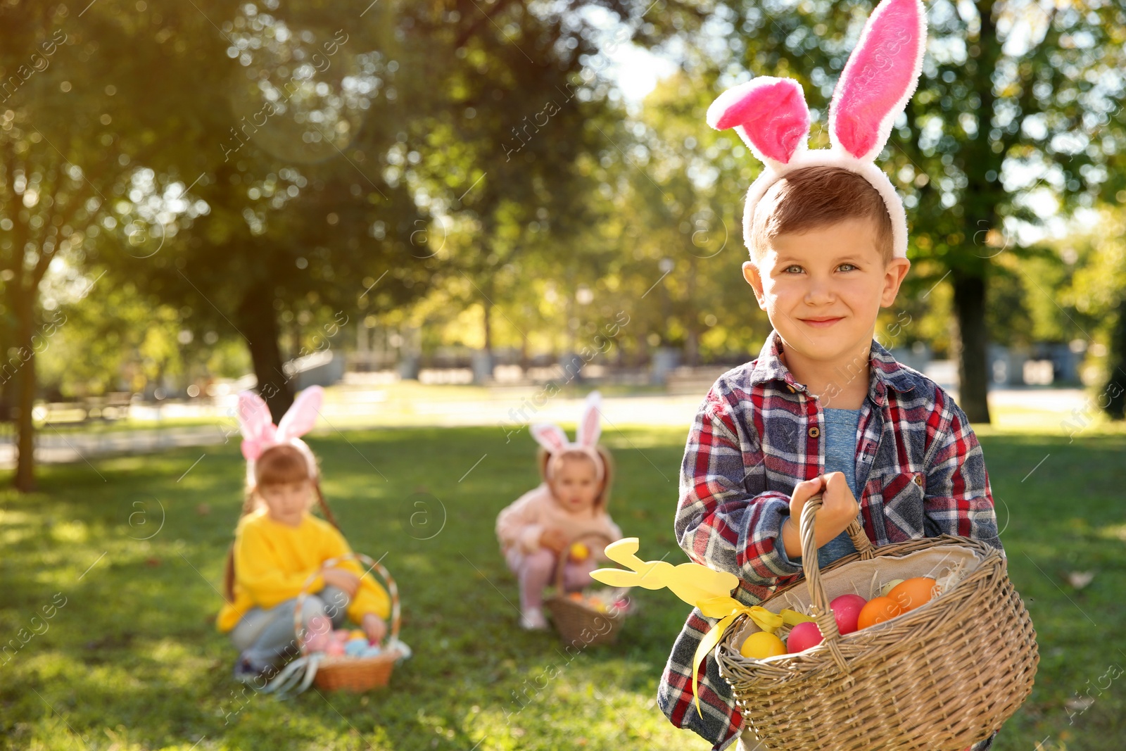 Photo of Cute little boy with bunny ears and basket of Easter eggs in park. Space for text