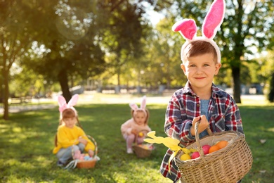 Photo of Cute little boy with bunny ears and basket of Easter eggs in park. Space for text