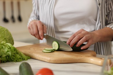 Woman cutting cucumber at white marble table, closeup