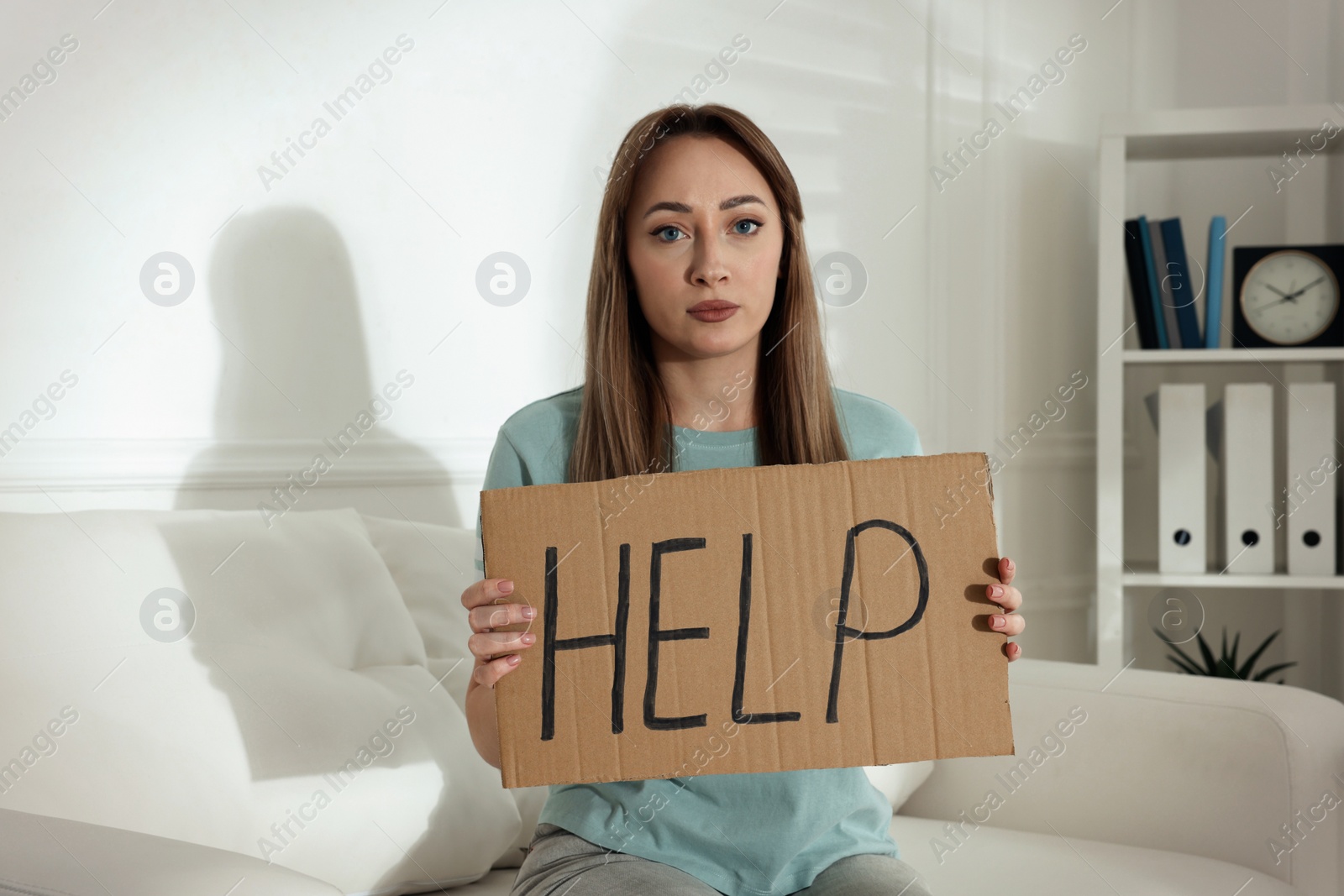 Photo of Unhappy young woman with HELP sign on sofa indoors