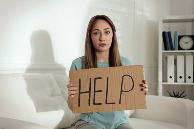 Unhappy young woman with HELP sign on sofa indoors