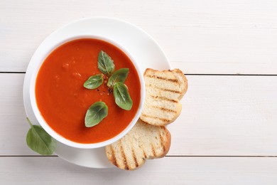 Delicious tomato cream soup in bowl with pieces of grilled bread on white wooden table, top view. Space for text