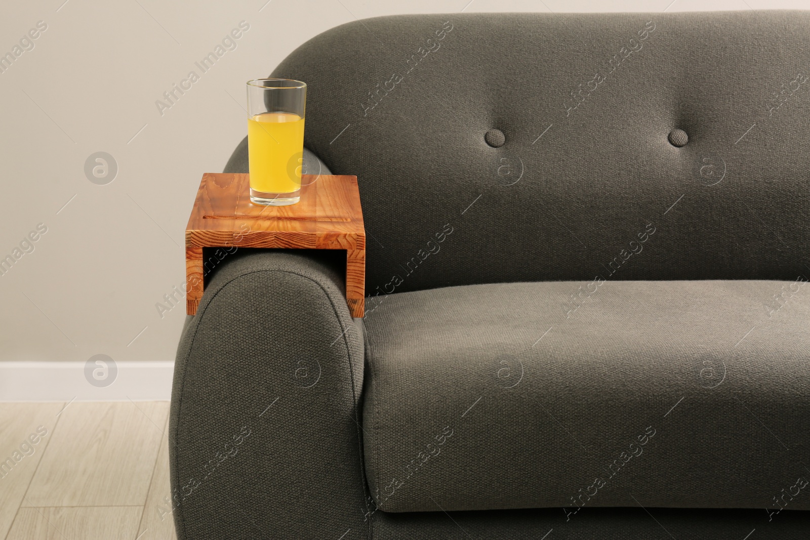 Photo of Glass of juice on sofa with wooden armrest table in room. Interior element