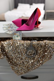 Prostitution concept. High heeled shoes, dollar banknotes, sequin dress and handcuffs on white chest of drawers indoors