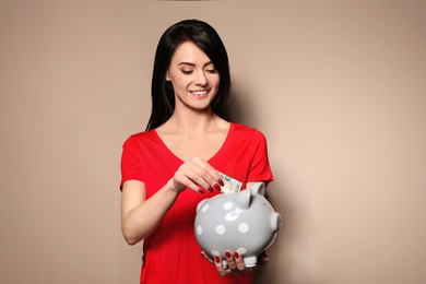 Beautiful woman putting money into piggy bank on color background