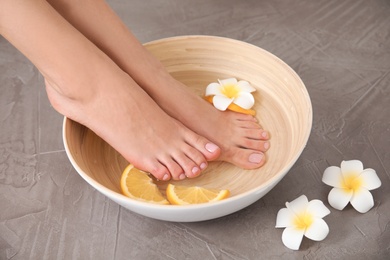 Woman soaking her feet in bowl with water, orange slices and flower on grey background, closeup. Spa treatment