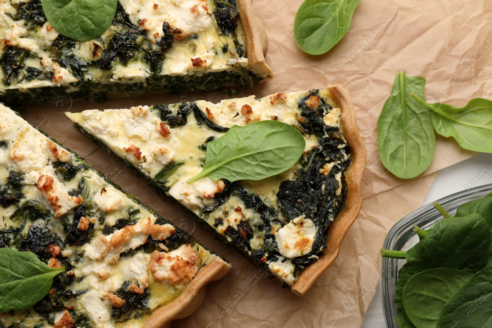 Photo of Delicious homemade quiche and fresh spinach leaves on parchment paper, flat lay
