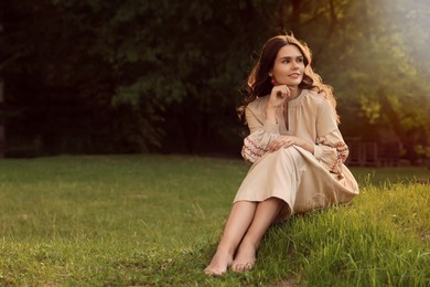 Photo of Beautiful woman in embroidered dress sitting on green grass outdoors, space for text. Ukrainian national clothes