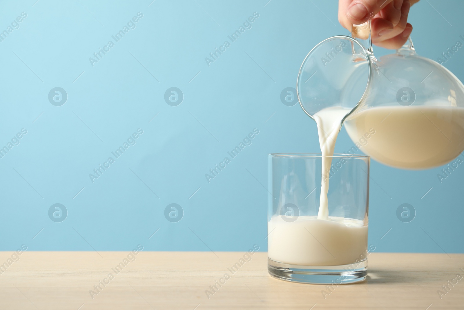 Photo of Woman pouring milk into glass at wooden table against light blue background, closeup. Space for text