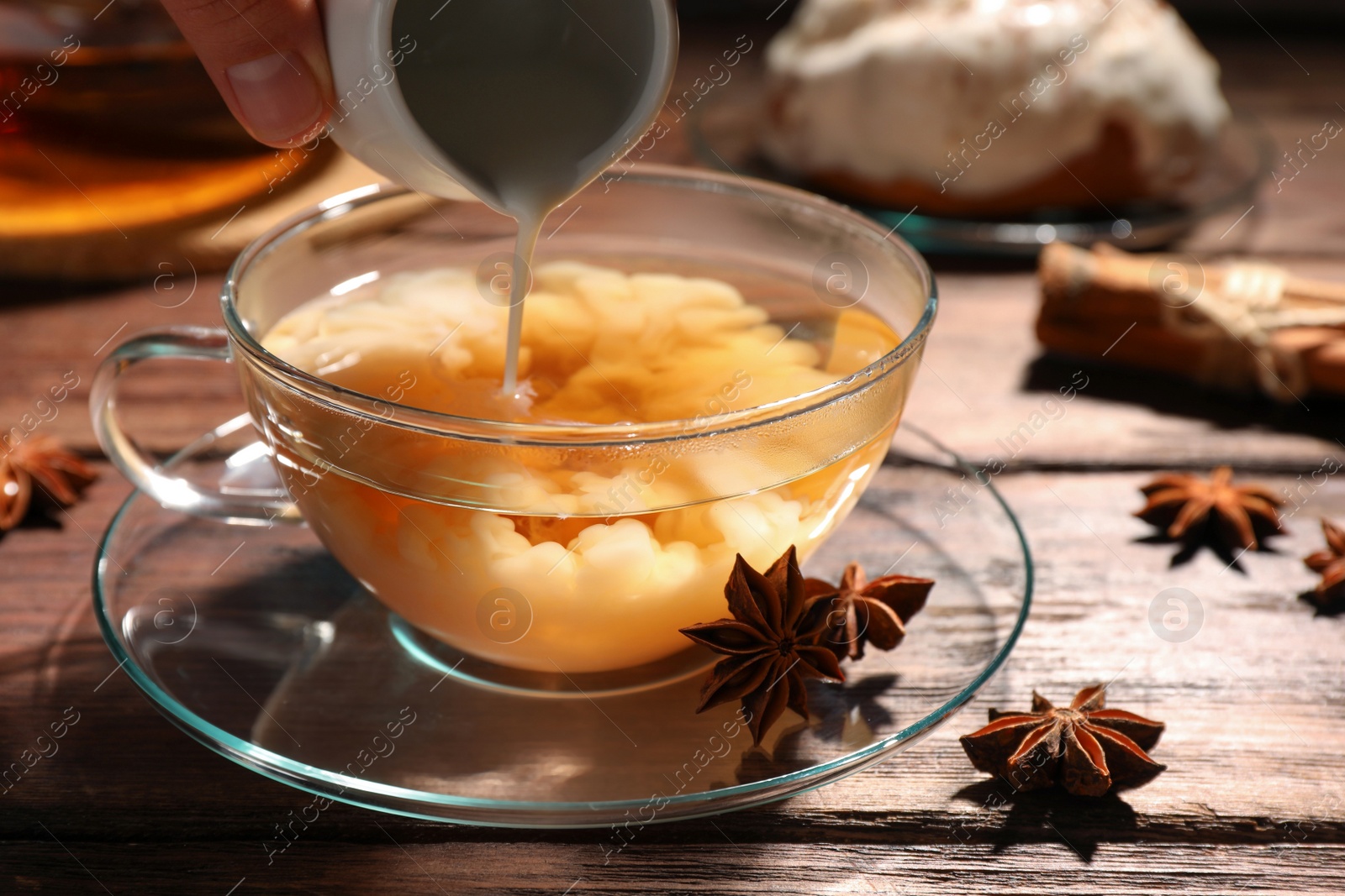 Photo of Woman pouring milk into glass cup of tea with anise stars at wooden table, closeup