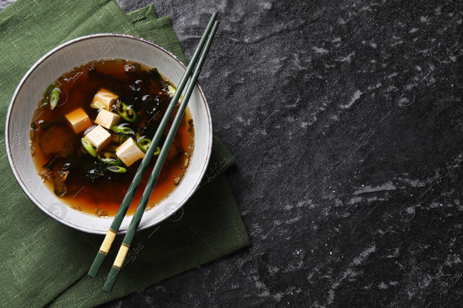 Photo of Bowl of delicious miso soup with tofu and chopsticks on black textured table, top view. Space for text