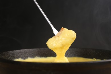Photo of Dipping piece of bread into fondue pot with tasty melted cheese against dark gray background, closeup