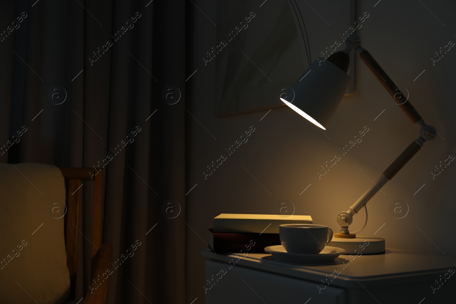 Photo of Stylish lamp, cup of drink and books on table in dark room. Interior design