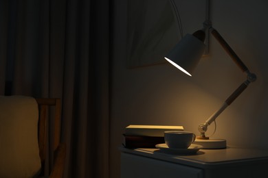 Stylish lamp, cup of drink and books on table in dark room. Interior design