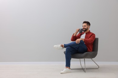 Handsome man with cup of drink sitting in armchair near light grey wall indoors, space for text