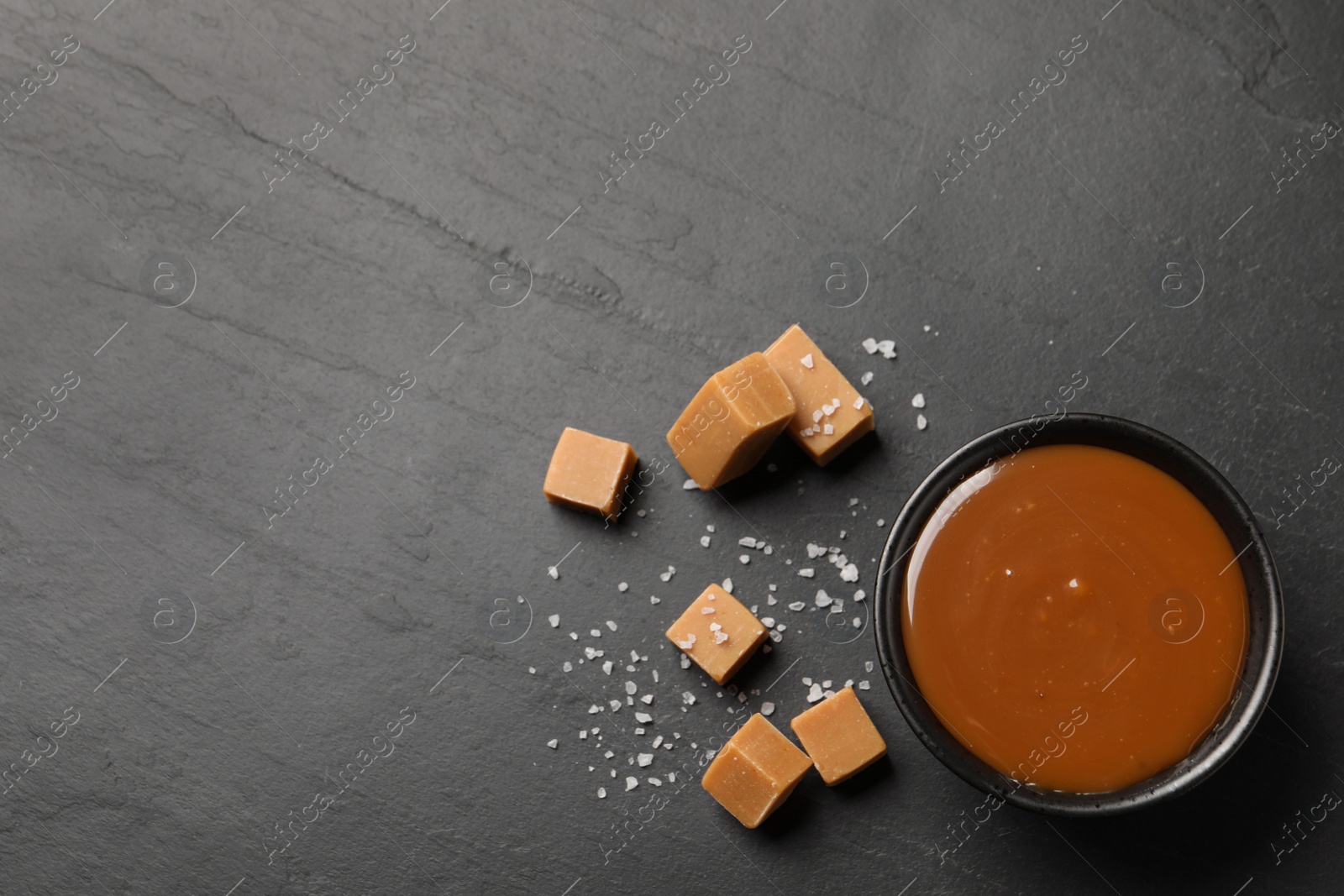 Photo of Yummy salted caramel in bowl and candies on black table, flat lay. Space for text