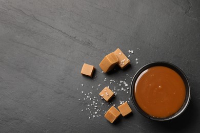 Photo of Yummy salted caramel in bowl and candies on black table, flat lay. Space for text