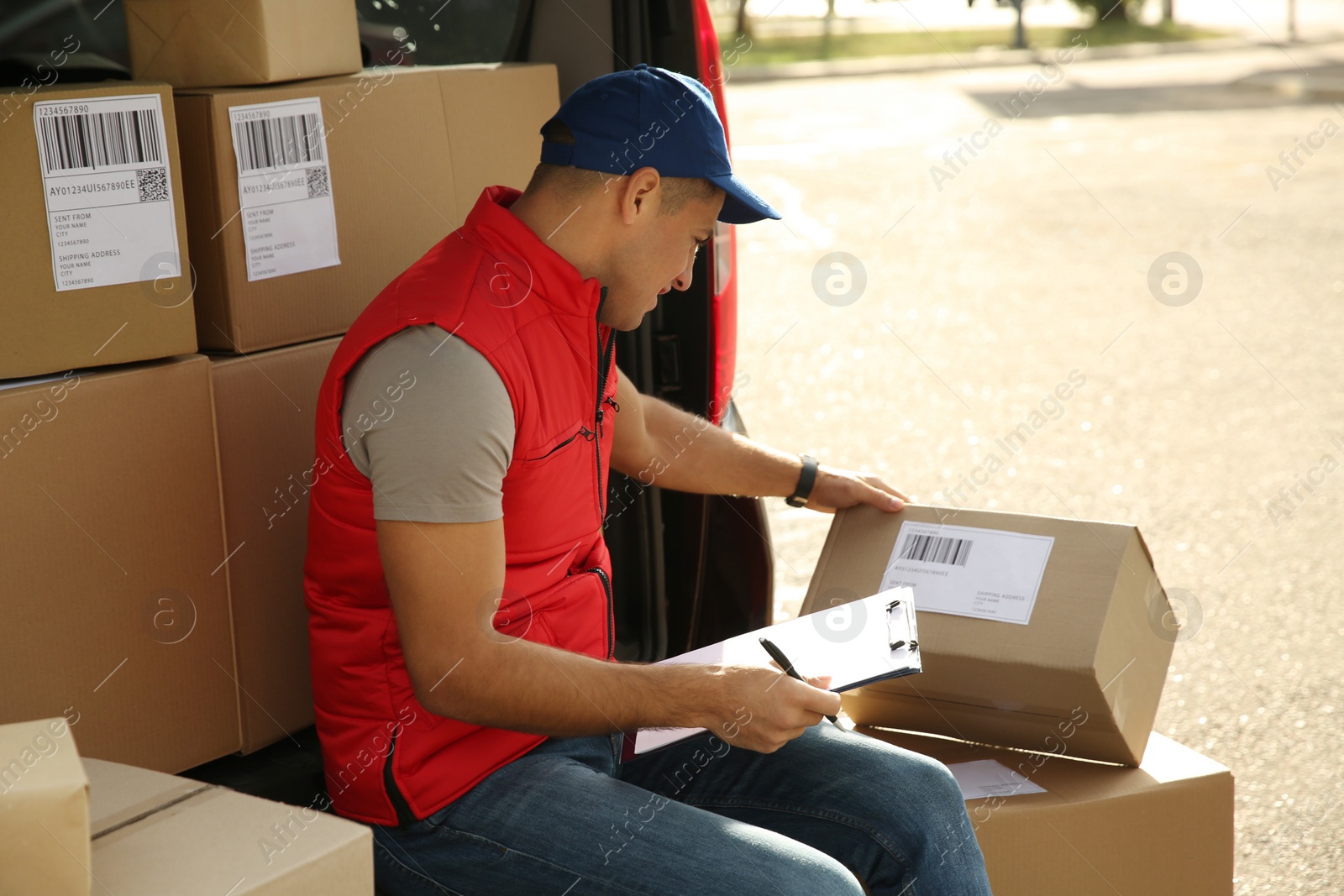 Photo of Courier with clipboard checking packages near car outdoors