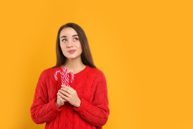 Young woman in red sweater holding candy canes on yellow background, space for text. Celebrating Christmas