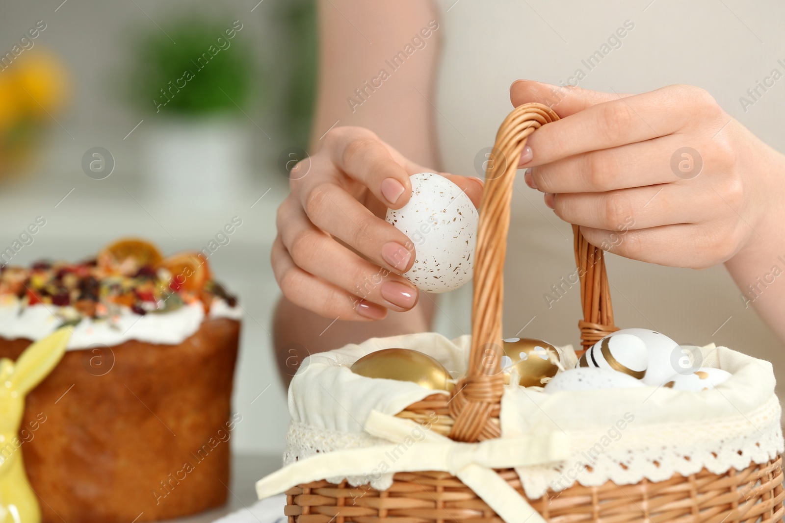 Photo of Closeup of woman putting painted egg into Easter basket near cake