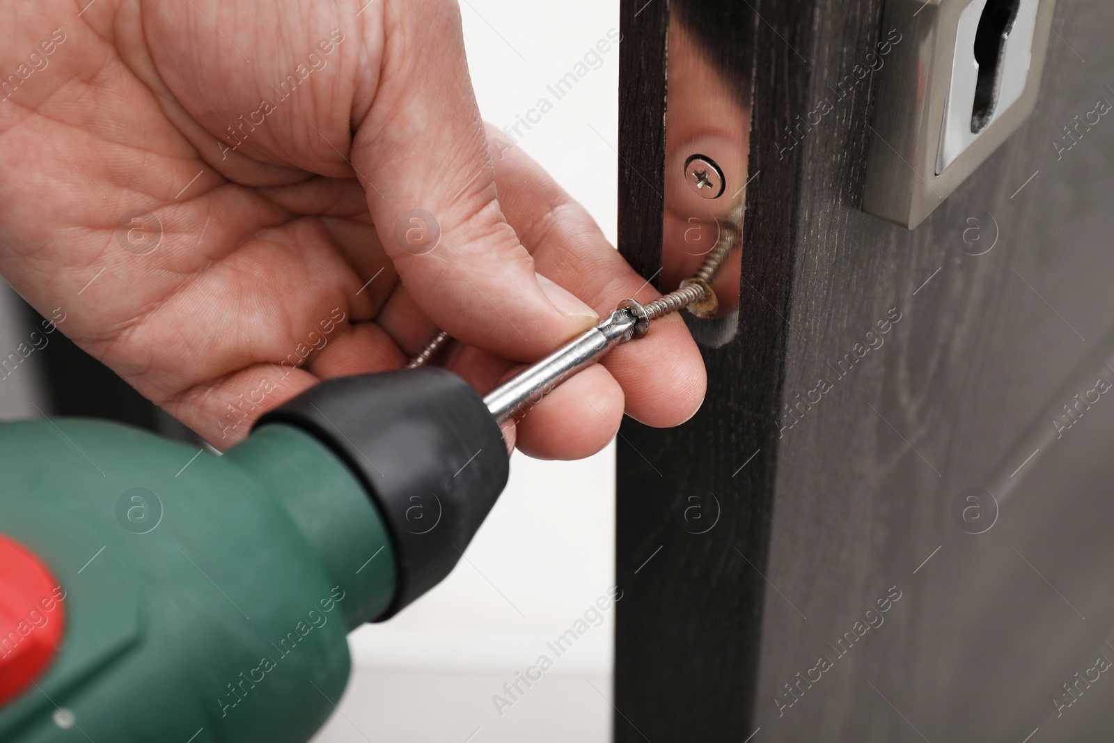 Photo of Handyman with electric screwdriver repairing door handle indoors, closeup