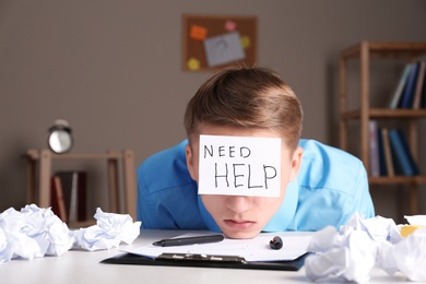 Photo of Young man with note NEED HELP on forehead at workplace