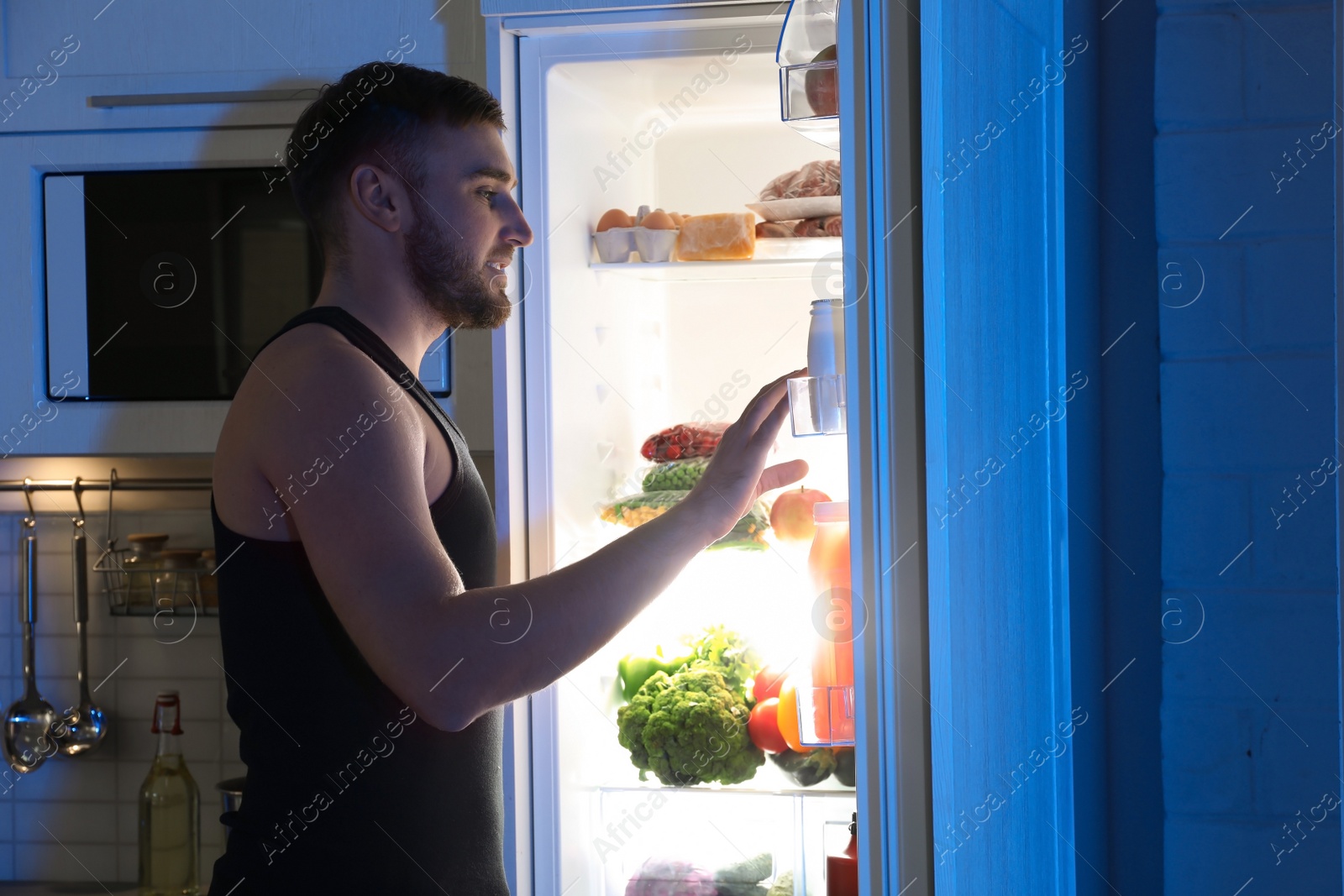 Photo of Man looking into refrigerator and choosing products in kitchen at night