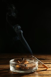 Photo of Glass ashtray with stubs and smoldering cigarette on wooden table against black background. Space for text