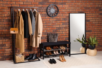 Photo of Stylish hallway with coat rack, mirror and shoe storage bench near brick wall. Interior design