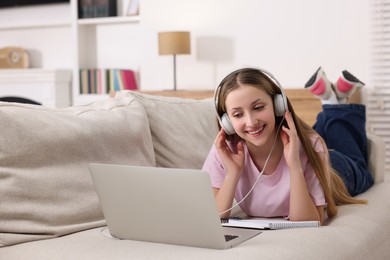 Photo of Online learning. Smiling teenage girl in headphones near laptop on sofa at home