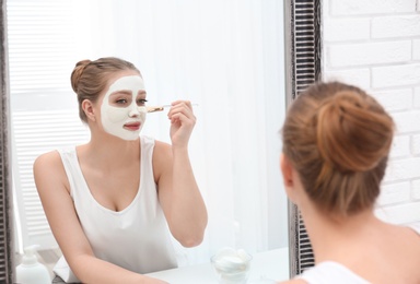 Beautiful woman applying homemade clay mask on her face at mirror indoors