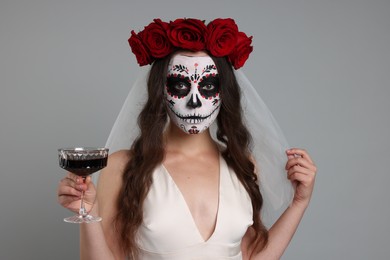 Photo of Young woman in scary bride costume with sugar skull makeup, flower crown and glass of wine on light grey background. Halloween celebration