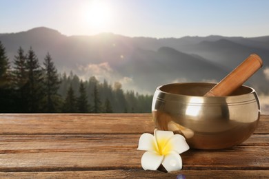 Image of Golden singing bowl, mallet and flower on wooden table against mountain landscape, space for text