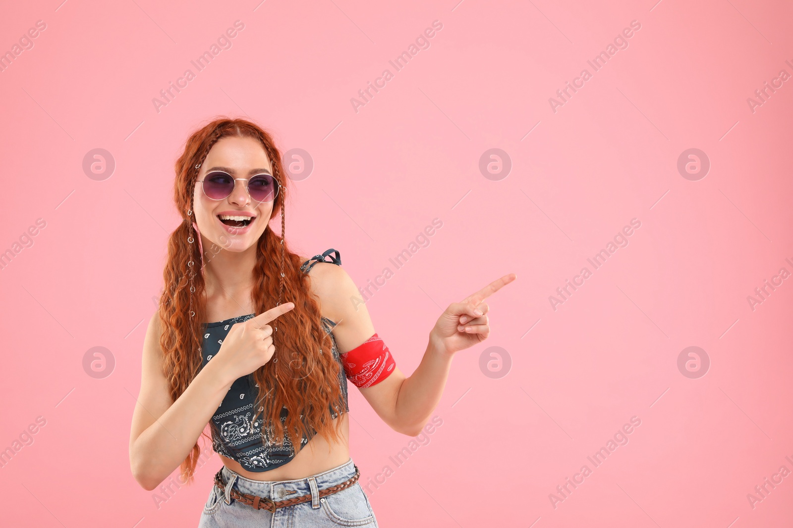 Photo of Stylish young hippie woman in sunglasses pointing at something on pink background, space for text