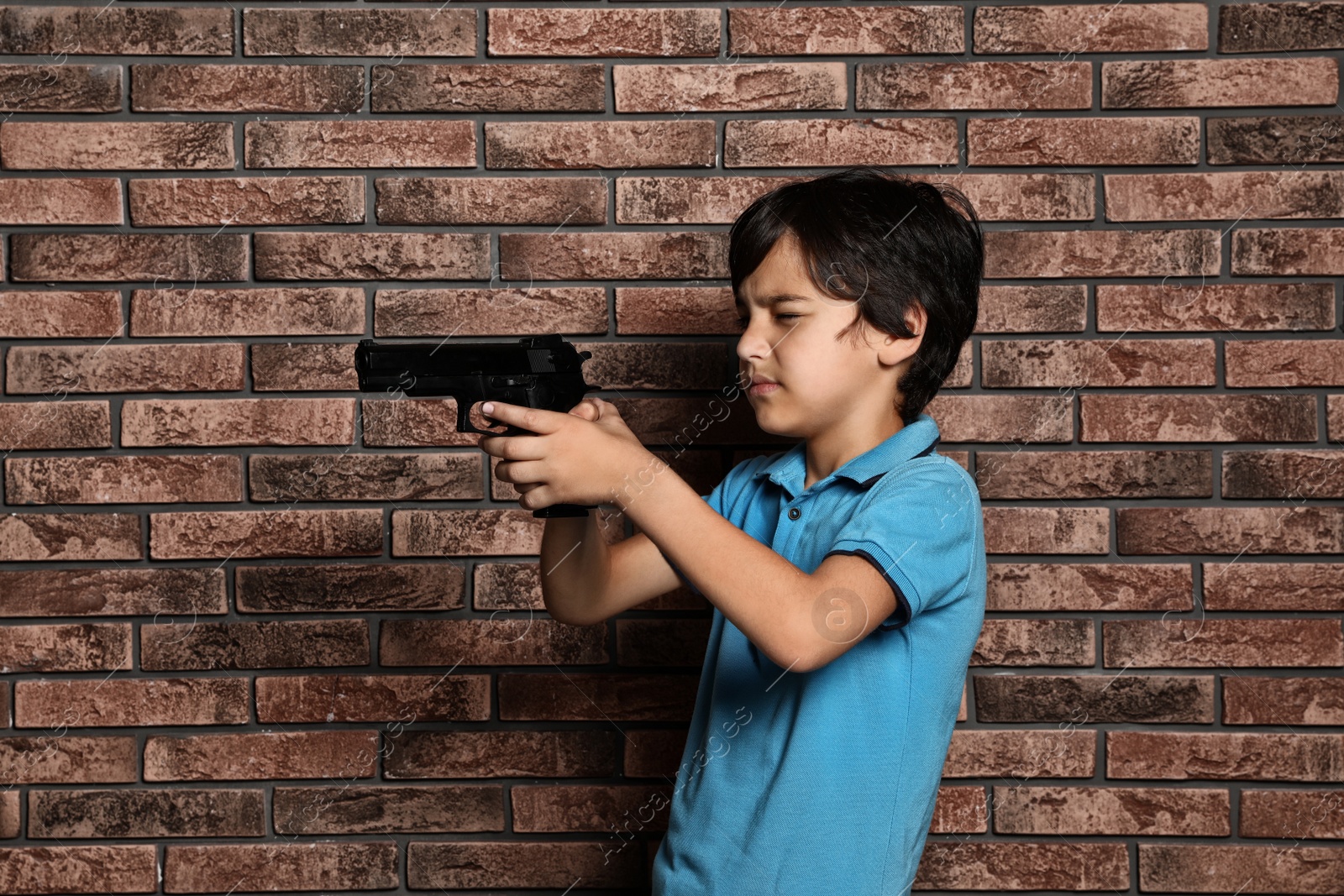 Photo of Little child playing with gun against brick wall. Dangerous game