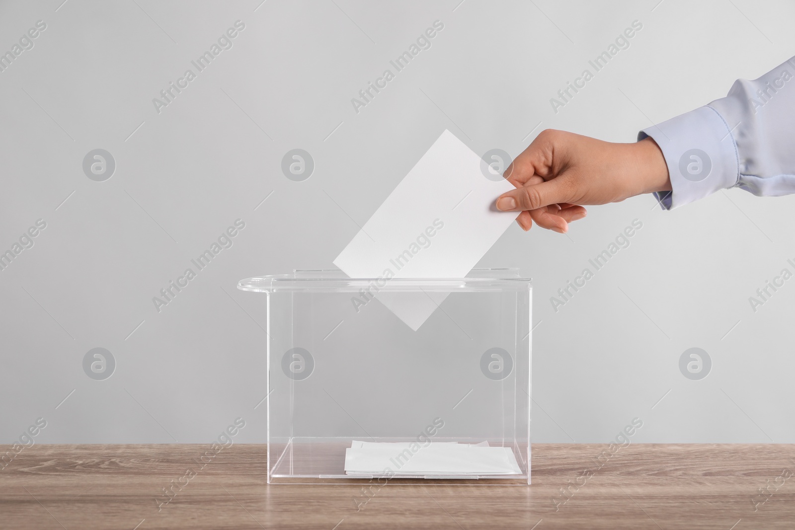 Photo of Woman putting her vote into ballot box on wooden table against light grey background, closeup