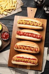 Photo of Delicious hot dogs with mustard, ketchup and potato fries on black wooden table, flat lay