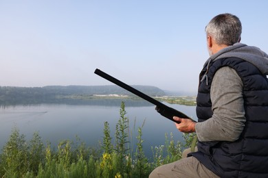 Photo of Man with hunting rifle sitting on wooden bench near lake outdoors. Space for text