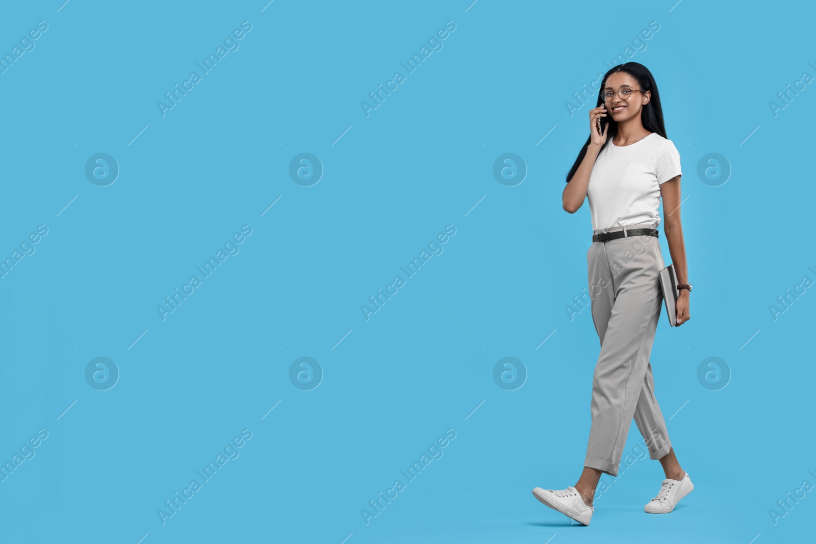 Photo of Beautiful secretary with laptop talking on phone against light blue background, space for text