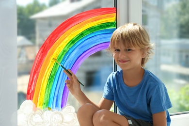 Photo of Little boy drawing rainbow on window indoors. Stay at home concept