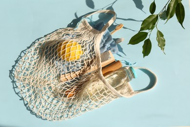 Photo of Fishnet bag with different items and leaves on light blue background, top view. Conscious consumption