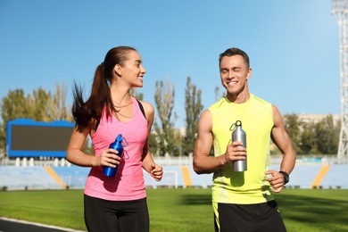 Photo of Young sporty couple running with bottles of water at stadium on sunny day