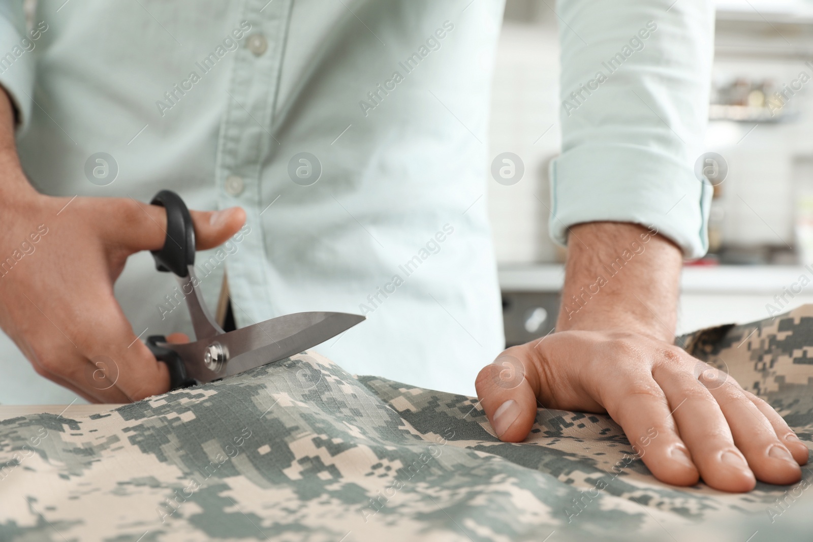 Photo of Professional tailor cutting camouflage fabric with scissors in workshop, closeup