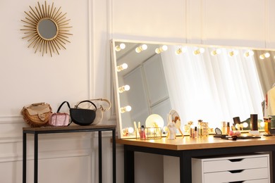 Photo of Makeup room. Stylish mirror with light bulbs, beauty products and bags on wooden table