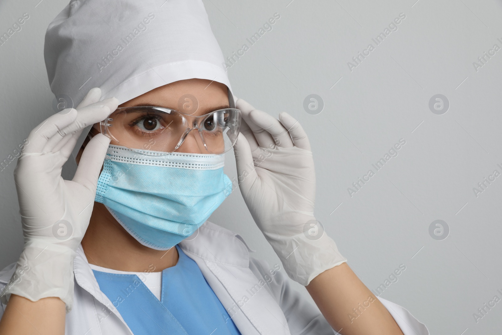 Photo of Doctor in protective mask and medical gloves putting on glasses against light grey background